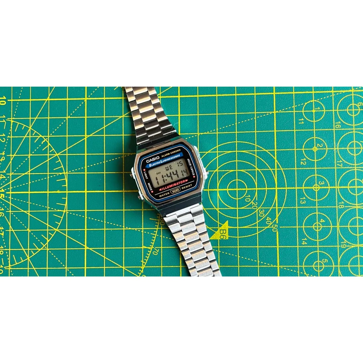 Casio A168WA-1 vintage digital watch with retro LCD display and stainless steel bracelet shown in a flat lay arrangement on a cutting mat surface.