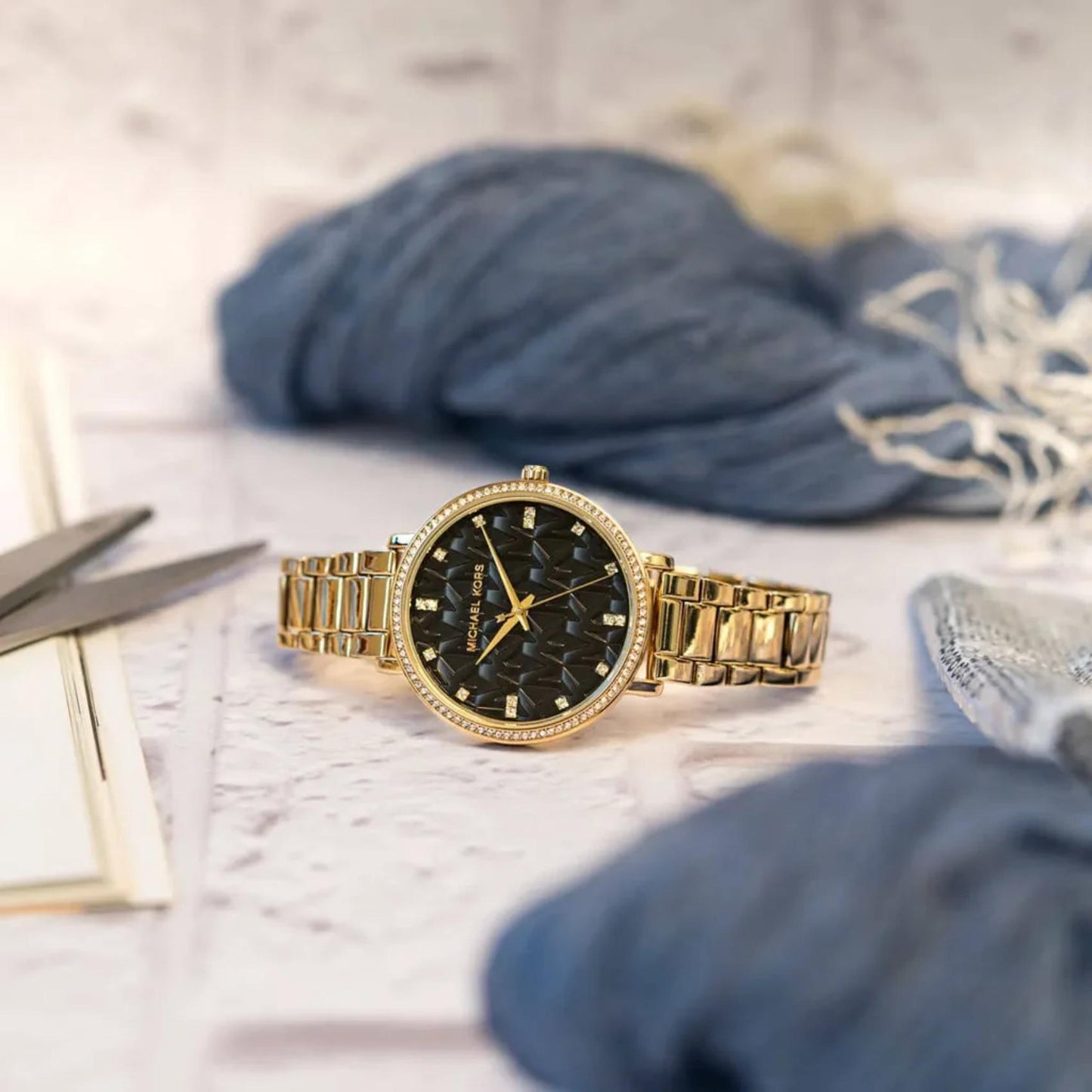 Michael Kors women’s watch with black MK logo dial, crystal bezel and gold-tone stainless steel bracelet placed in lifestyle setting.