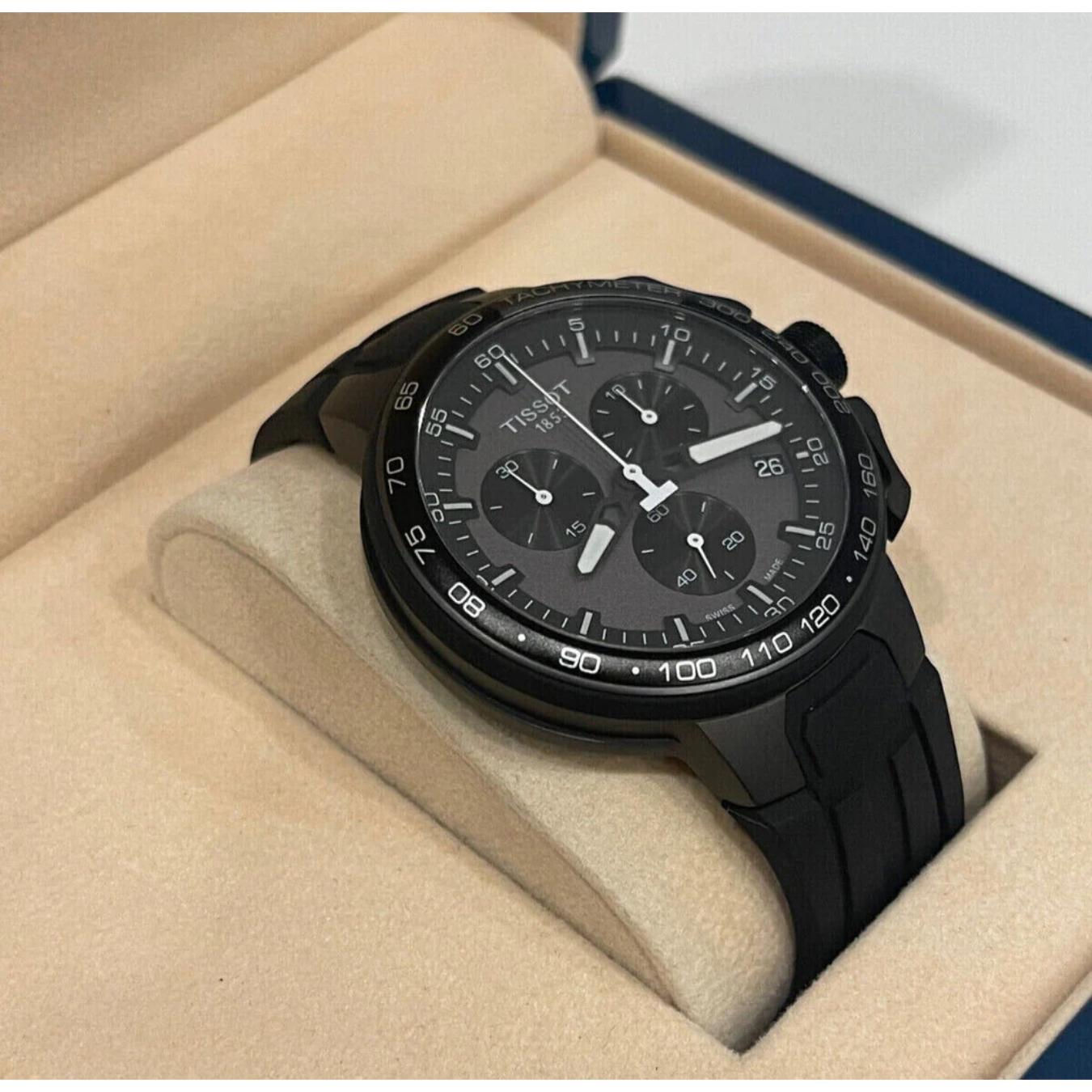 Reloj cronógrafo negro Tissot T-Race Cycling T111.417.37.441.03 con correa de caucho, apoyado en un cojín beige dentro de su estuche de presentación abierto