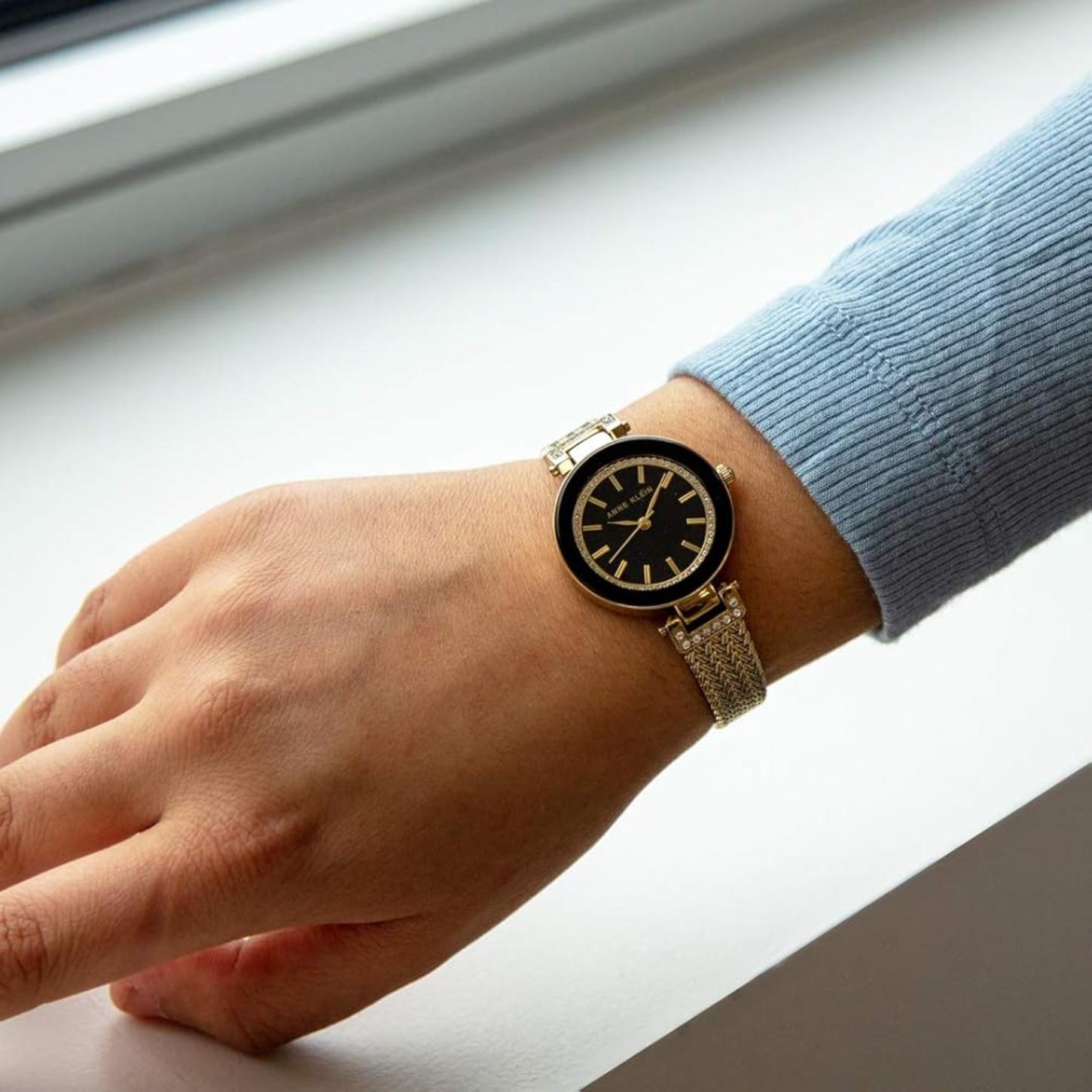 Anne Klein AK/1906BKGB with black dial, crystal-accented bezel, and gold-tone mesh bracelet worn on the wrist in an elegant everyday style.