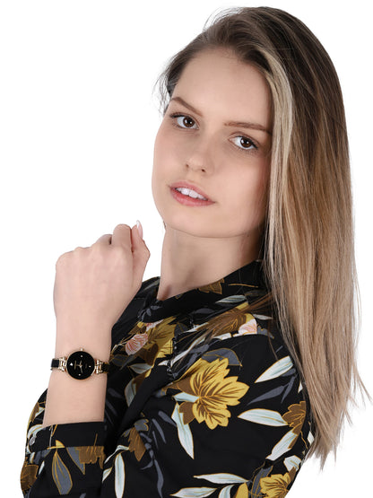 Reloj Anne Klein AK/1980BKGB en muñeca de mujer, esfera negra y brazalete dorado combinados con blusa floral oscura.
