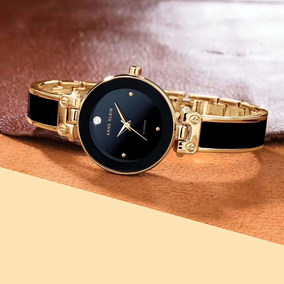 Reloj Anne Klein AK/1980BKGB apoyado sobre superficie, esfera negra con diamante y brazalete negro y dorado en primer plano