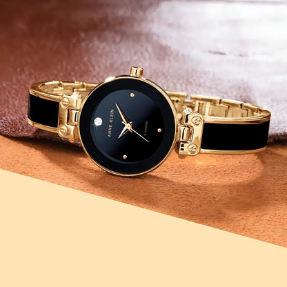 Reloj Anne Klein AK/1980BKGB apoyado sobre superficie, esfera negra con diamante y brazalete negro y dorado en primer plano