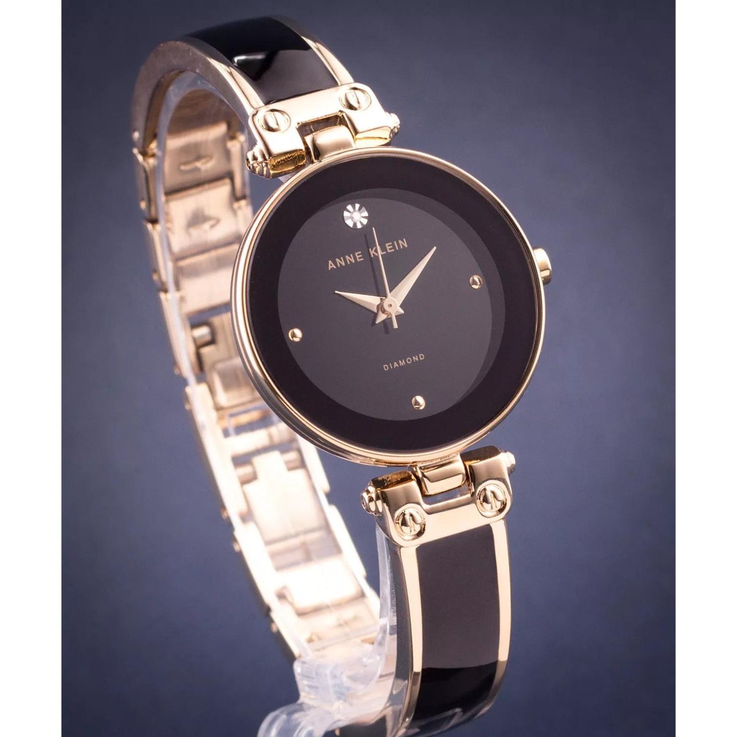 Reloj Anne Klein AK/1980BKGB en vista tres cuartos, esfera negra con diamante y brazalete negro y dorado sobre fondo neutro