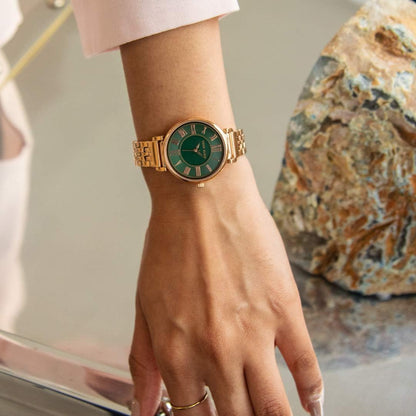 Reloj Anne Klein AK/2158GNRG en muñeca de mujer, caja y brazalete oro rosa con esfera verde junto a roca decorativa
