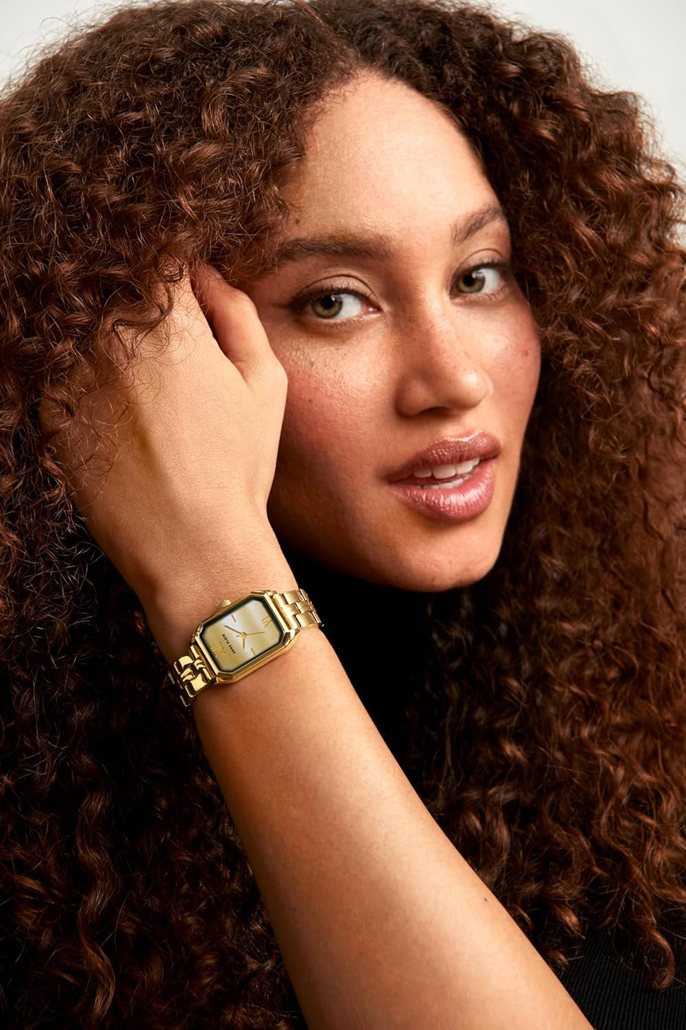 Reloj Anne Klein AK/3774CHGB en muñeca de mujer, brazalete y caja rectangulares dorados sobre fondo de rizos oscuros.
