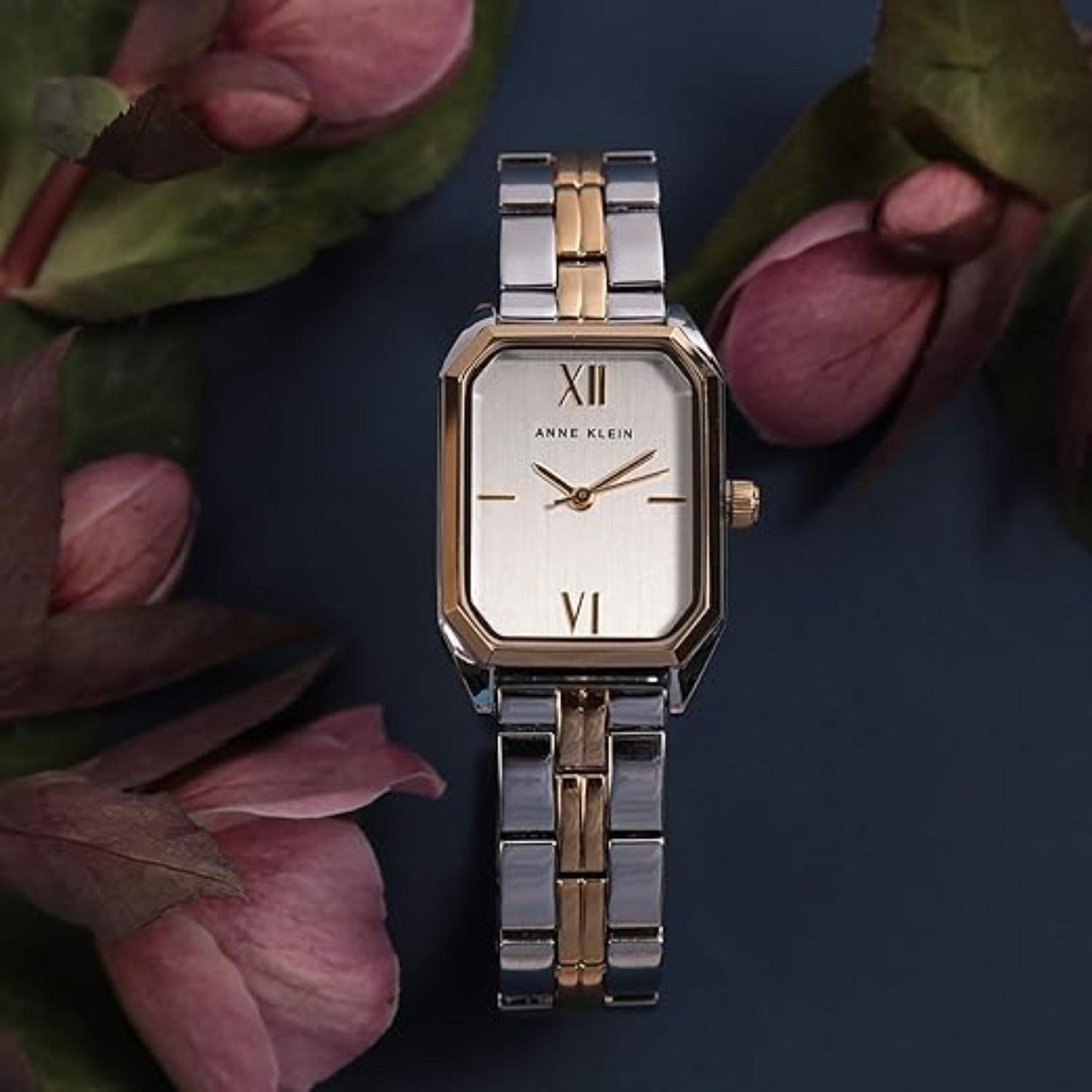 Anne Klein AK/3775SVTT women’s dress watch with silver rectangular dial, gold-tone bezel accents and two-tone bracelet displayed in elegant floral lifestyle setting.