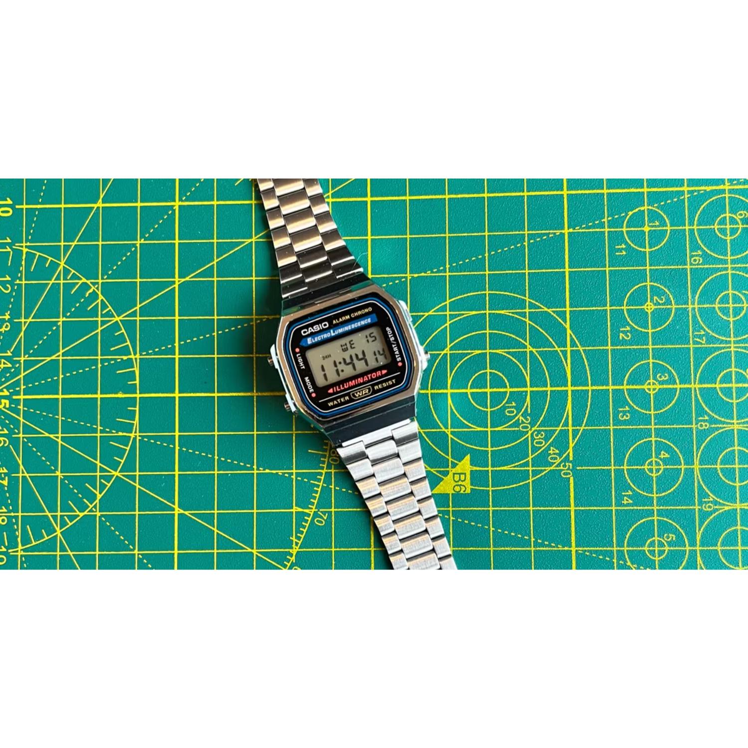 Casio A168WA-1 vintage digital watch with retro LCD display and stainless steel bracelet shown in a flat lay arrangement on a cutting mat surface.