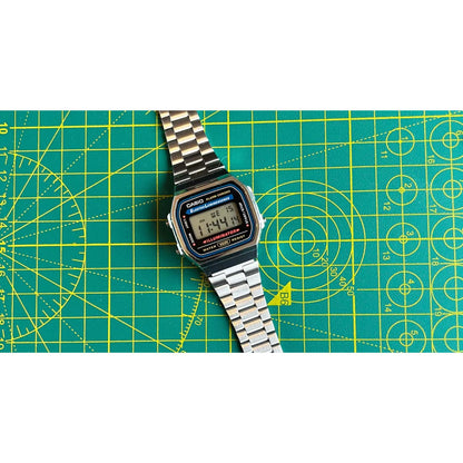Casio A168WA-1 vintage digital watch with retro LCD display and stainless steel bracelet shown in a flat lay arrangement on a cutting mat surface.