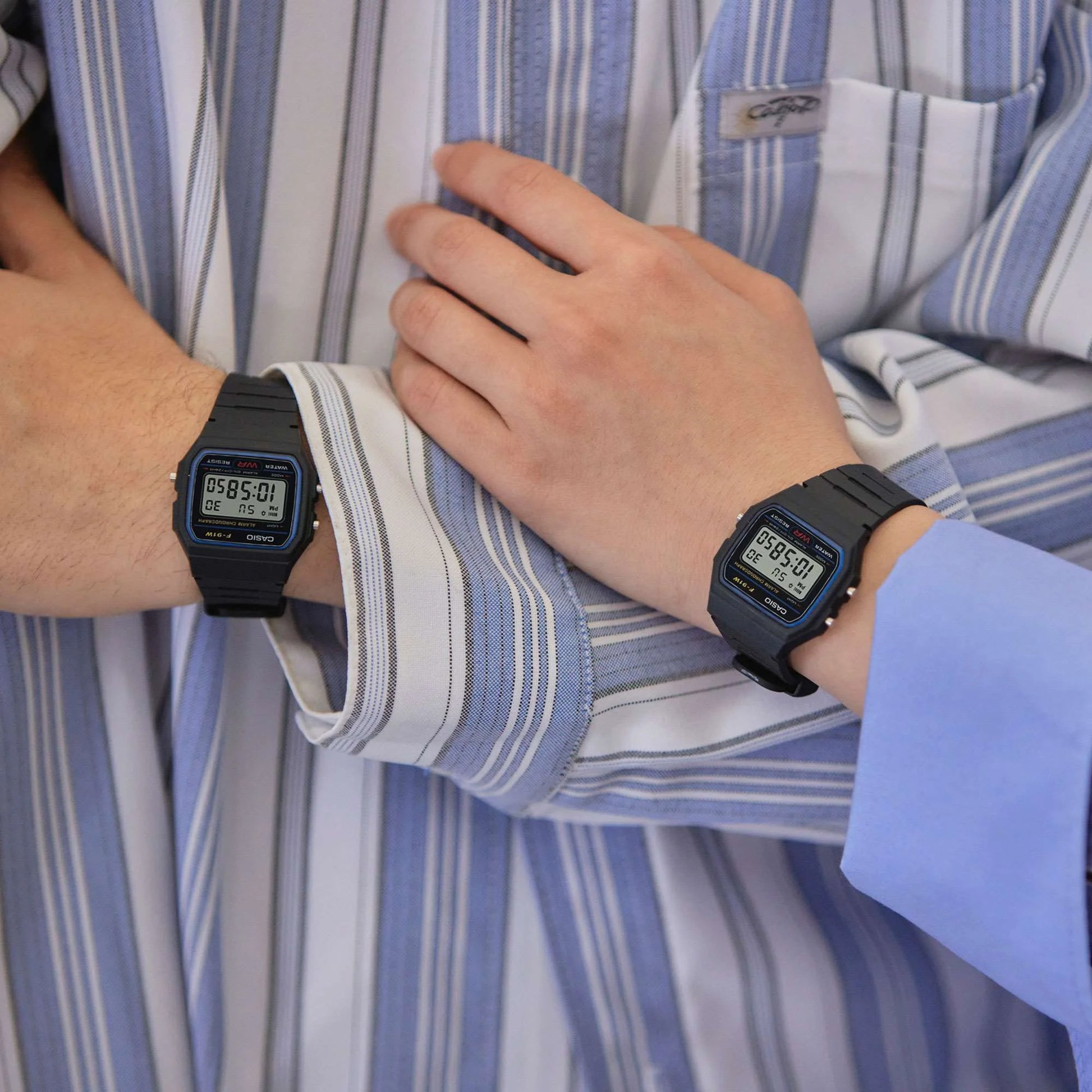 Dos personas muestran en la muñeca el reloj digital Casio F-91W, ambos con camisa de rayas.