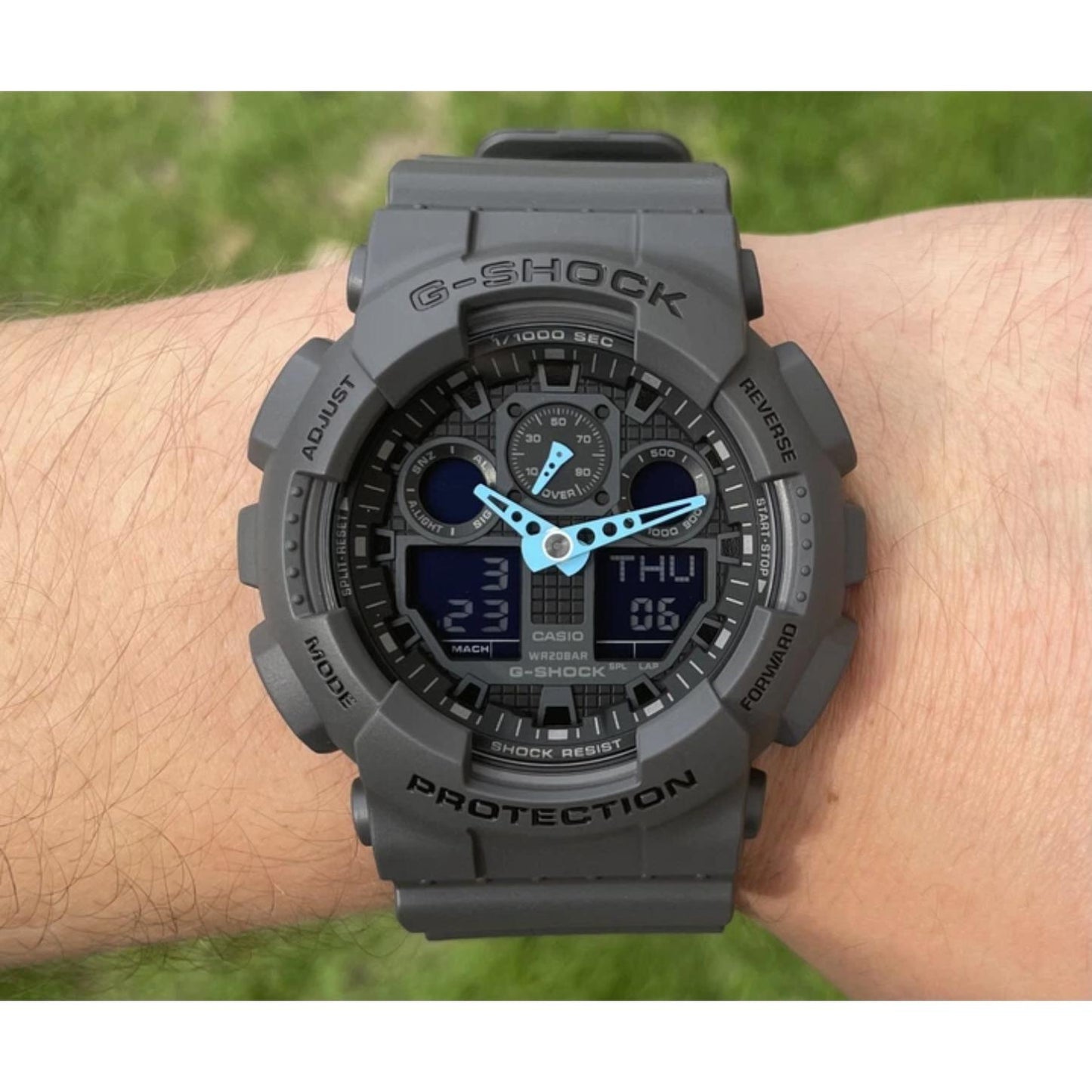 Casio G-Shock GA-100C-8A worn on the wrist outdoors, featuring a gray resin case and strap, dark analog-digital dial, and blue hands.