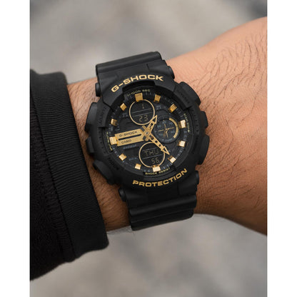 Casio G-Shock GA-100GB-1A watch with a black analog-digital dial, gold-tone hands and hour markers, black protective case, and matching black strap worn on the wrist.