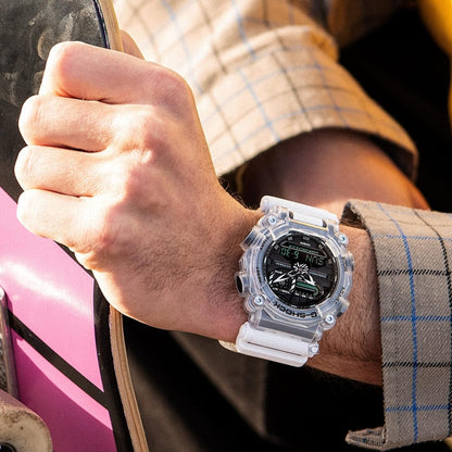 Casio G-Shock GA-900SKL-7ADR worn on the wrist with a skateboard, featuring a transparent resin case, white strap, black analog-digital dial, and rugged sport design.