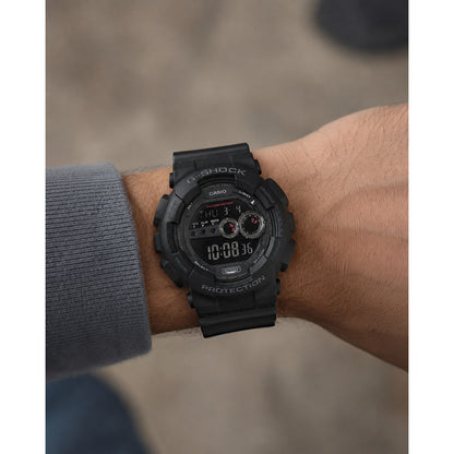 Casio G-Shock GD-100-1B men’s digital watch with a black negative display, red accent indicators, rugged black resin case, and matching black resin strap worn on the wrist.