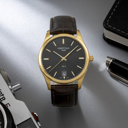 Certina DS-4 C022.610.36.081.00 men's watch on a flat surface alongside a camera, pen, and notebook, featuring a gold-tone case and gray dial