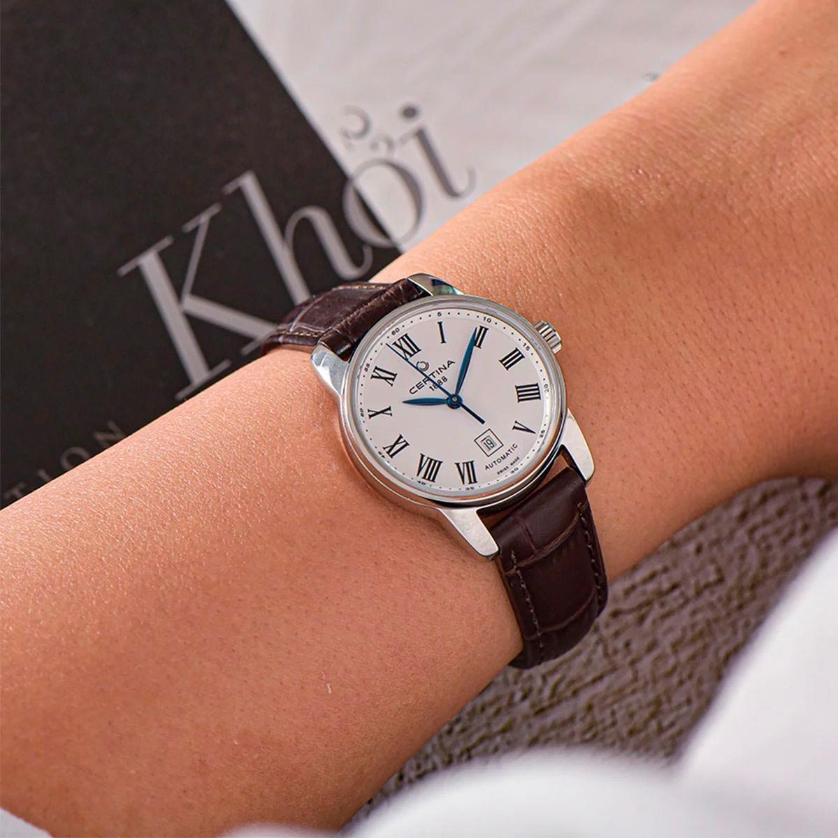 Certina DS Podium Lady C001.007.16.013.00 en muñeca con esfera blanca y números romanos, aguja azul central y correa de piel marrón.