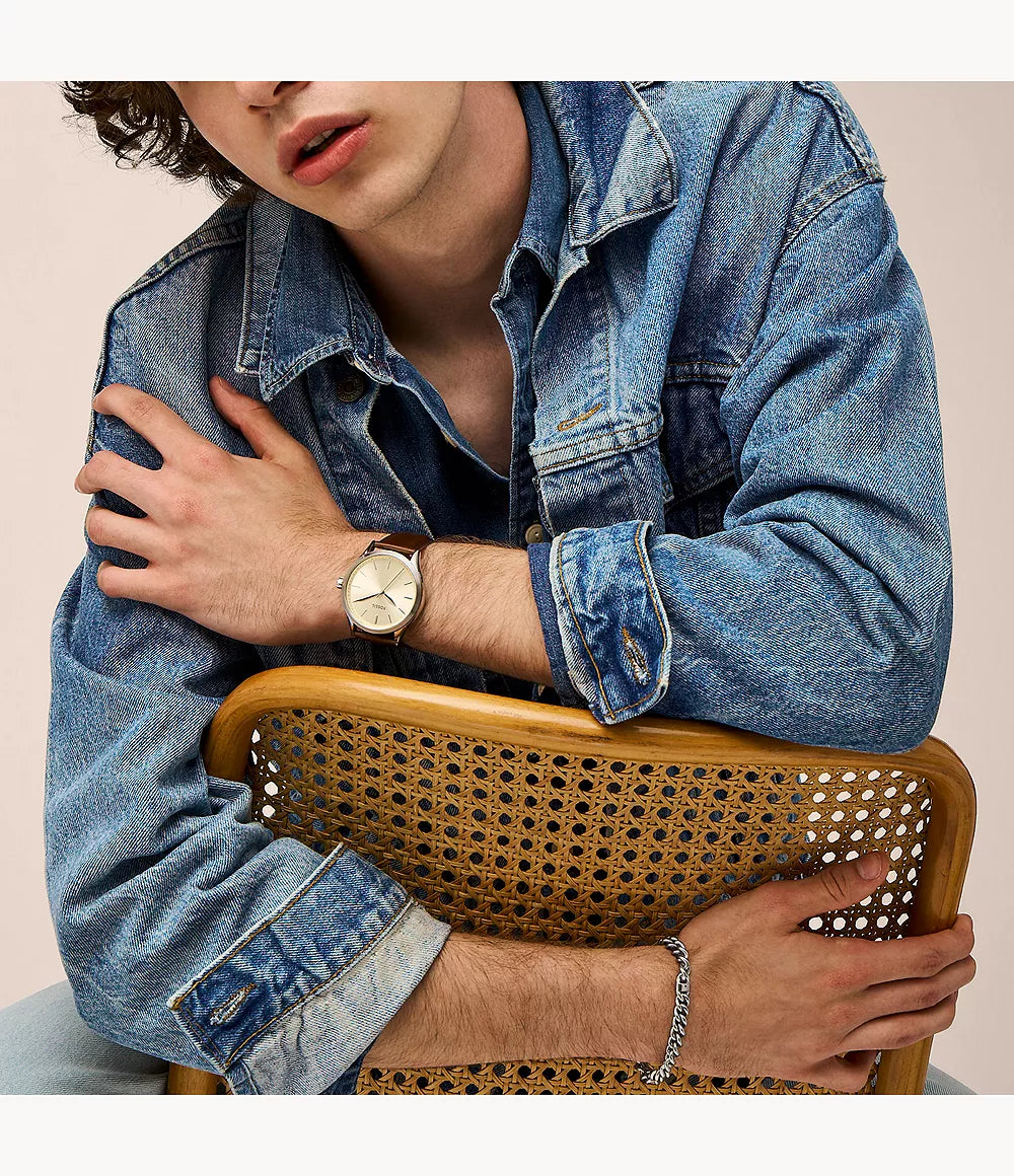 Reloj Fossil analógico con esfera dorada y correa de cuero marrón lucido en la muñeca con chaqueta vaquera.