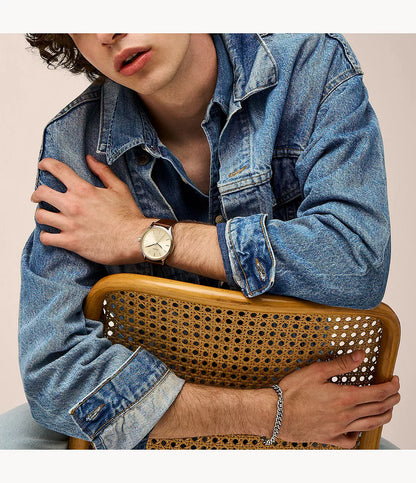 Reloj Fossil analógico con esfera dorada y correa de cuero marrón lucido en la muñeca con chaqueta vaquera.