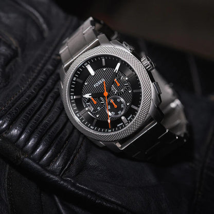 Fossil Machine FS6095 stainless steel chronograph watch with black textured dial and orange accents shown in a rugged lifestyle close-up view.