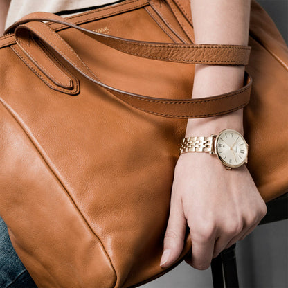 Reloj Fossil dorado de mujer con brazalete de acero, llevado en la muñeca junto a un bolso de piel marrón.