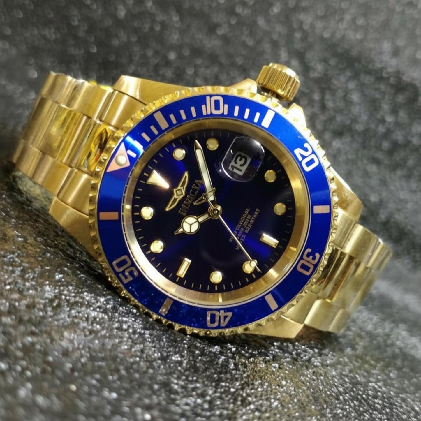 Reloj Invicta Pro Diver 26974 dorado con esfera y bisel azules visto en primer plano en posición inclinada sobre fondo brillante texturizado