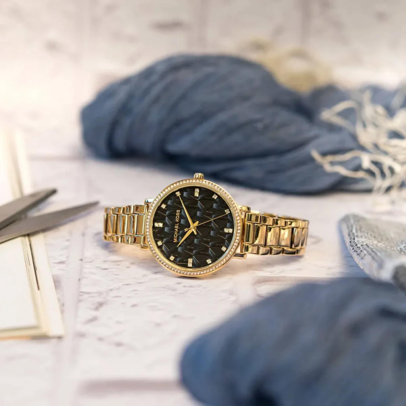 Michael Kors women’s watch with black MK logo dial, crystal bezel and gold-tone stainless steel bracelet placed in lifestyle setting.