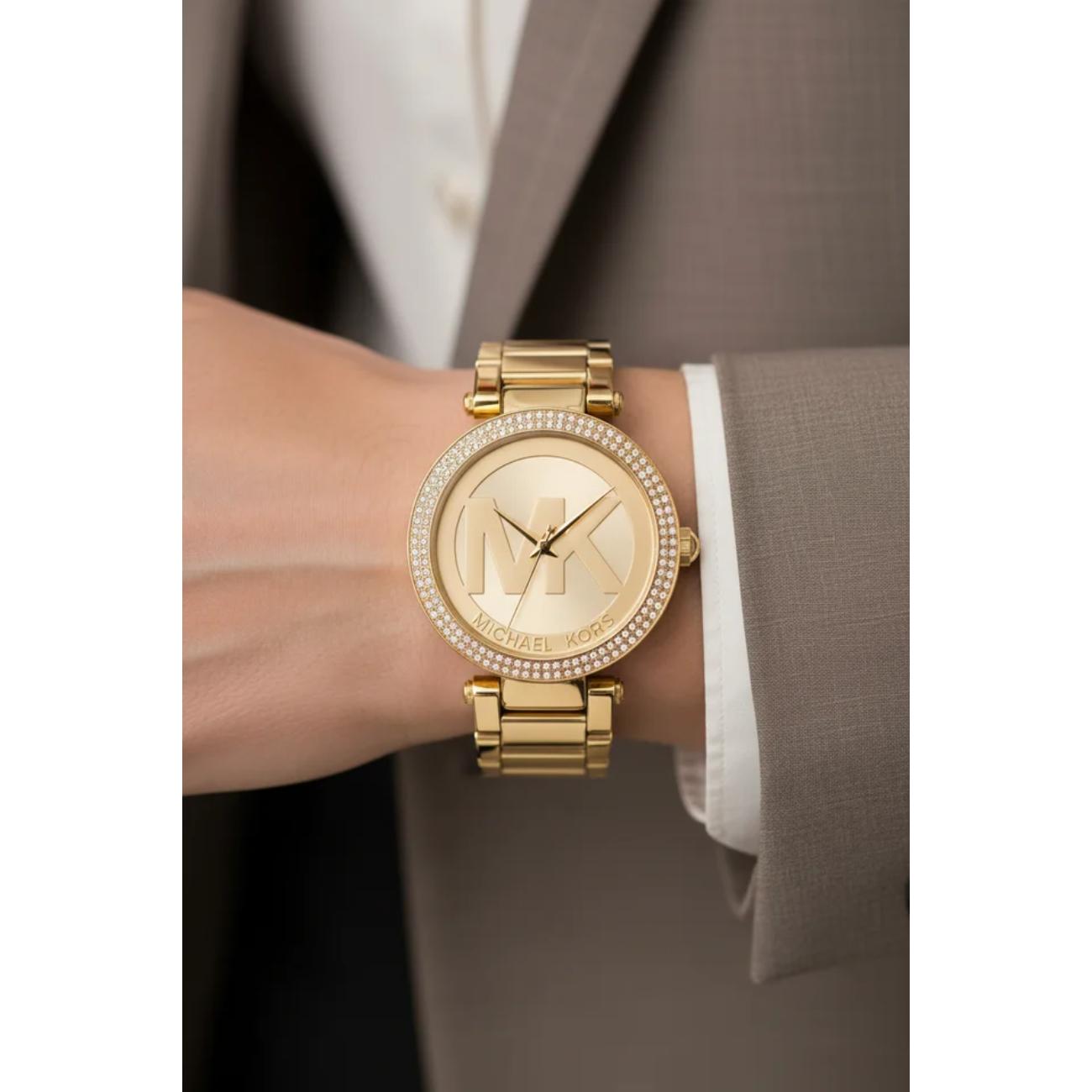 Michael Kors Parker MK5784 women’s gold-tone fashion watch with crystal-set bezel and oversized MK logo dial worn on the wrist with formal attire.