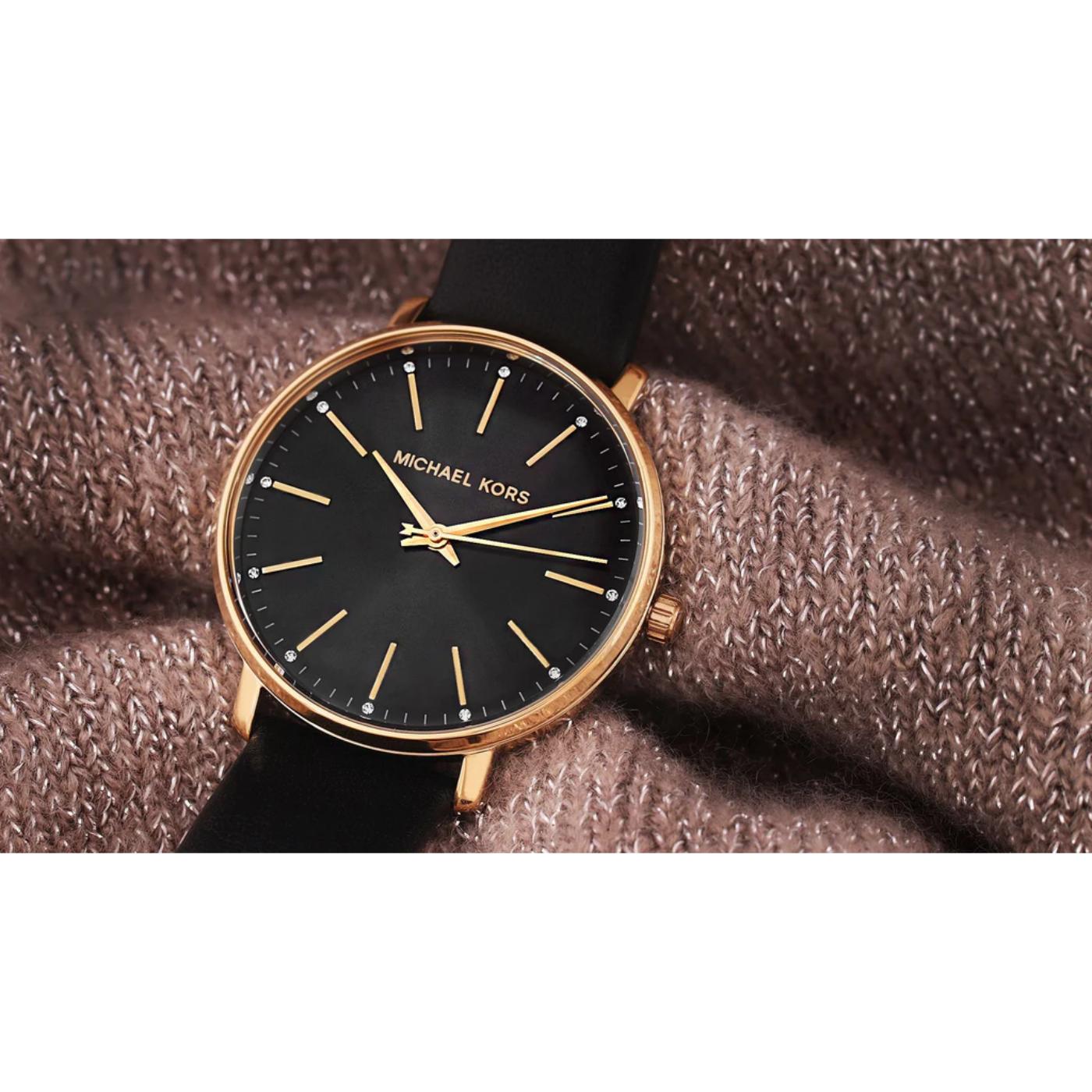 reloj Michael Kors Pyper MK2747 colocado sobre la muñeca, con esfera negra y caja dorada, fotografiado sobre un fondo textil marrón