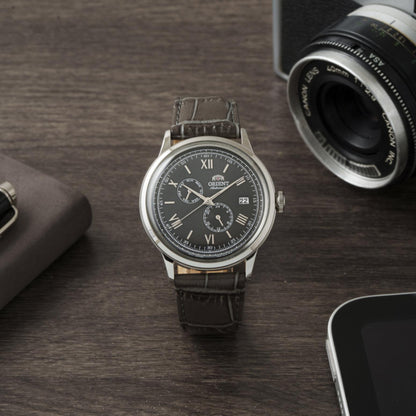 Orient Bambino Version 8 RA-AK0704N10B automatic dress watch with gray dial and dark crocodile-pattern leather strap displayed in desk lifestyle setting beside camera equipment.