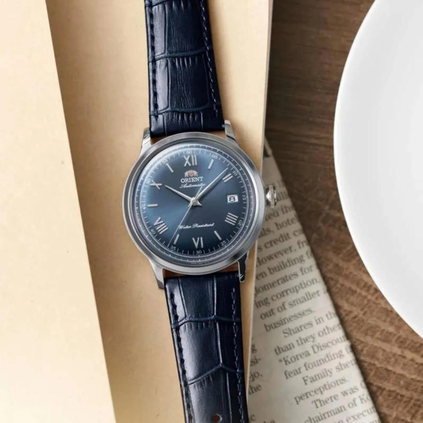 Orient RA-AC0024L30B automatic dress watch with blue sunburst dial and blue crocodile-pattern leather strap displayed on a table beside reading materials.