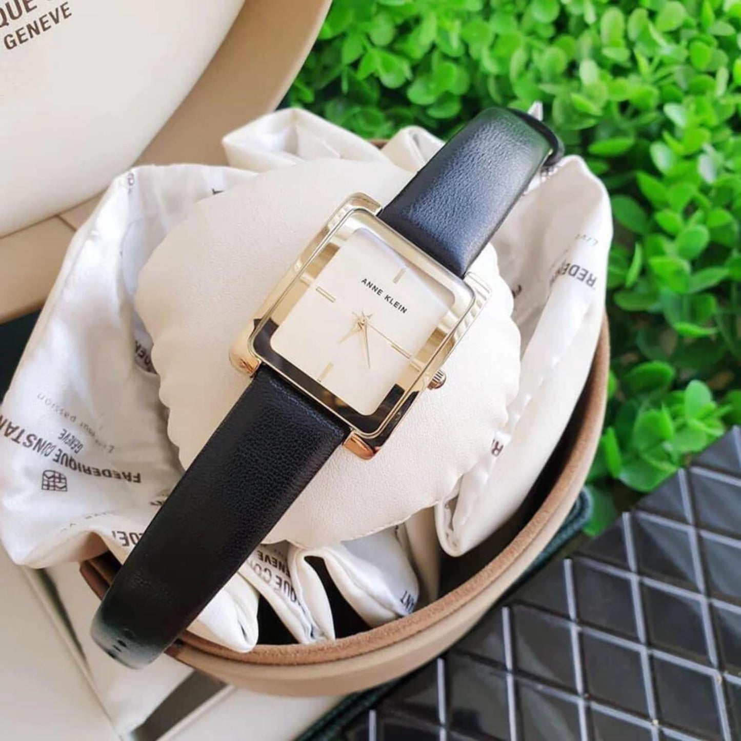 Reloj Anne Klein AK/2706CHBK con caja rectangular dorada y correa negra de piel, presentado sobre cojín en estuche, vista superior.