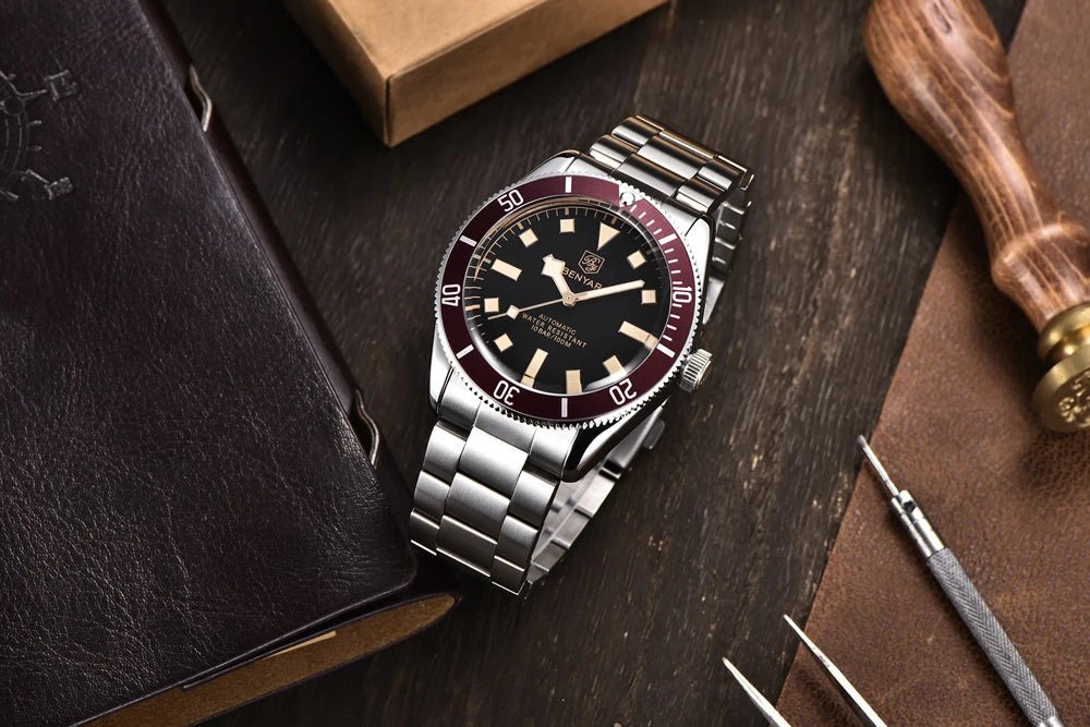 Reloj Benyar BB58 BY-5179M automático de acero, esfera negra y bisel rojo, junto a herramientas y libro, estilo relojero.