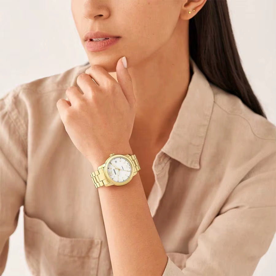 Reloj Fossil Heritage ME3226 automático dorado, puesto en la muñeca de mujer, camisa beige y pose elegante.