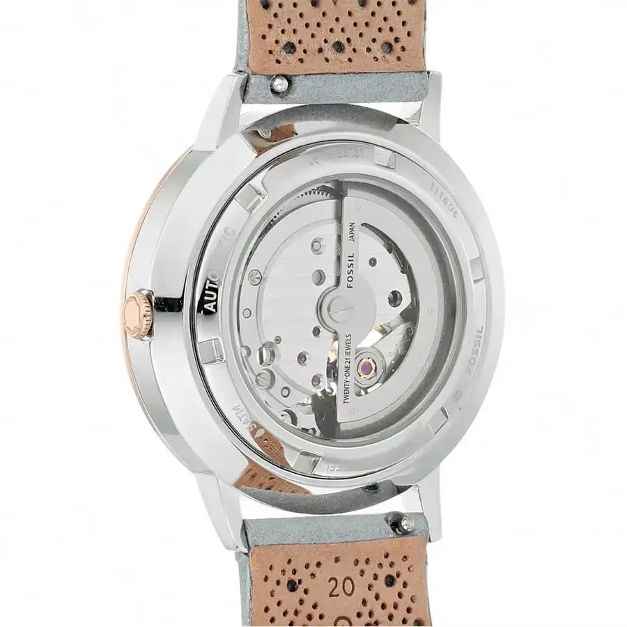 Vista trasera del reloj Fossil ME3131, fondo transparente mostrando la maquinaria automática, caja acero y detalles oro rosa, correa gris perforada.