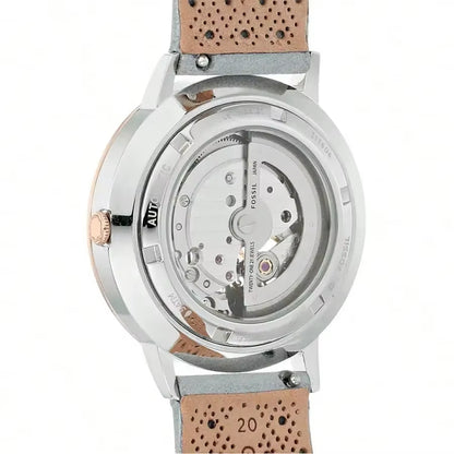 Vista trasera del reloj Fossil ME3131, fondo transparente mostrando la maquinaria automática, caja acero y detalles oro rosa, correa gris perforada.