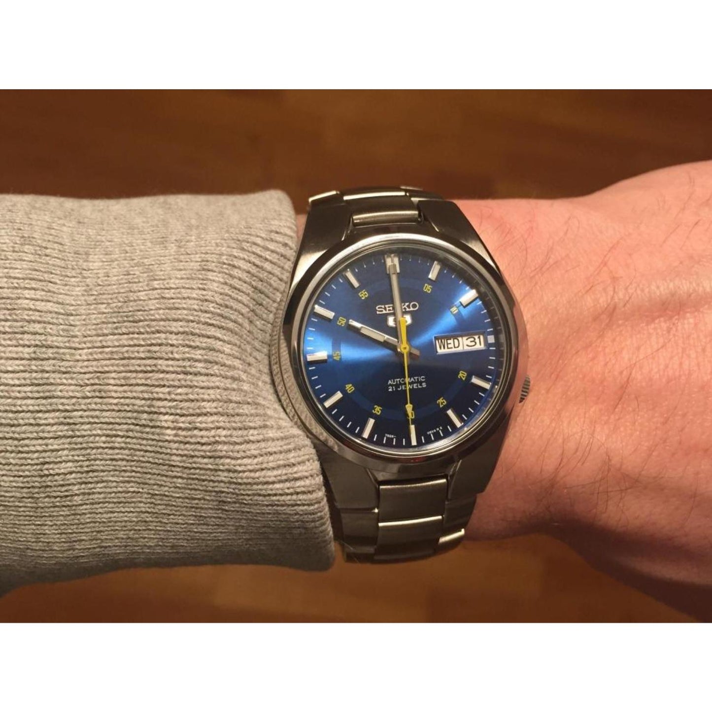 Muñeca masculina con un jersey gris claro luciendo el Seiko 5 SNK615K, destacando su esfera azul con efecto rayos de sol bajo una luz cálida y brazalete de acero integrado.