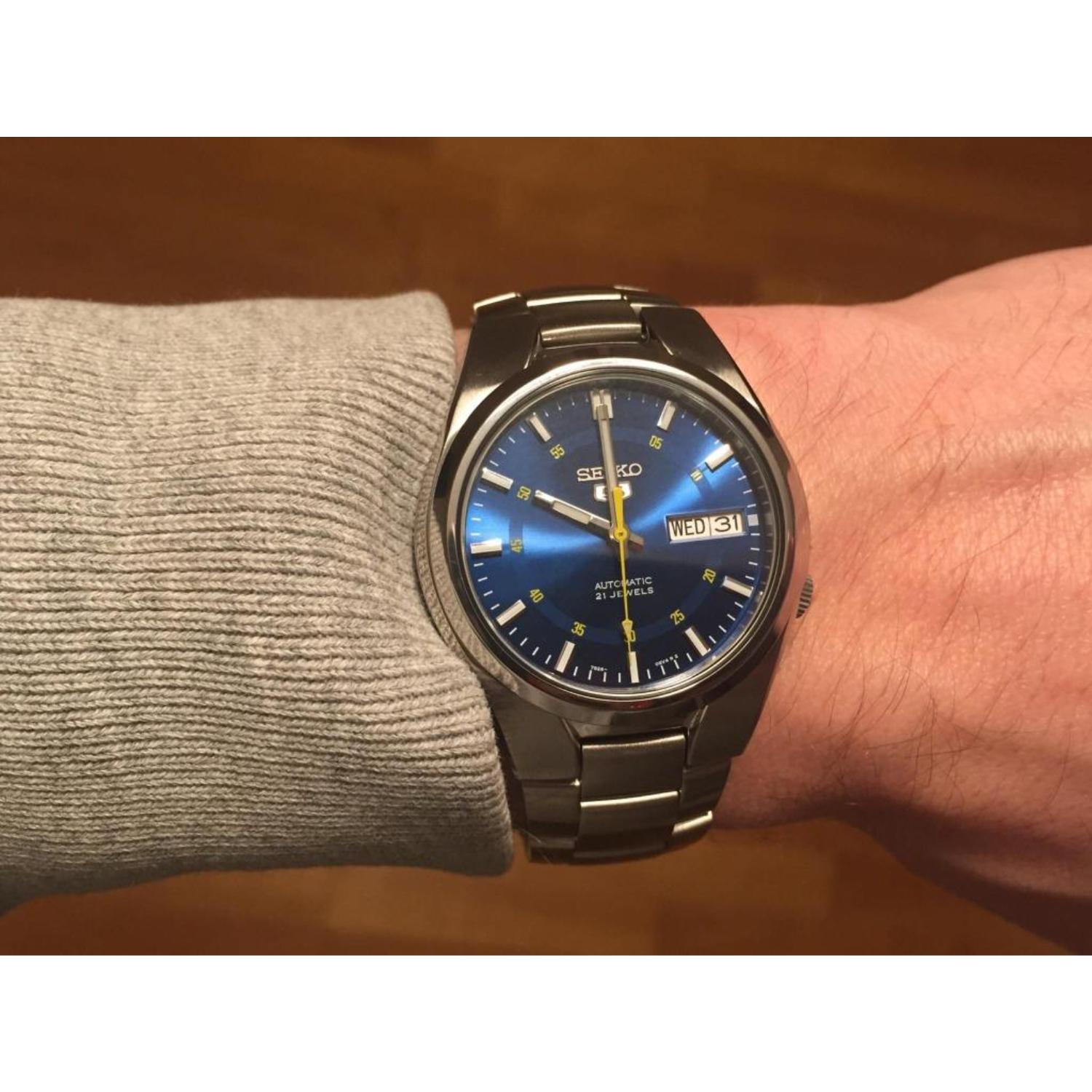 Muñeca masculina con un jersey gris claro luciendo el Seiko 5 SNK615K, destacando su esfera azul con efecto rayos de sol bajo una luz cálida y brazalete de acero integrado.
