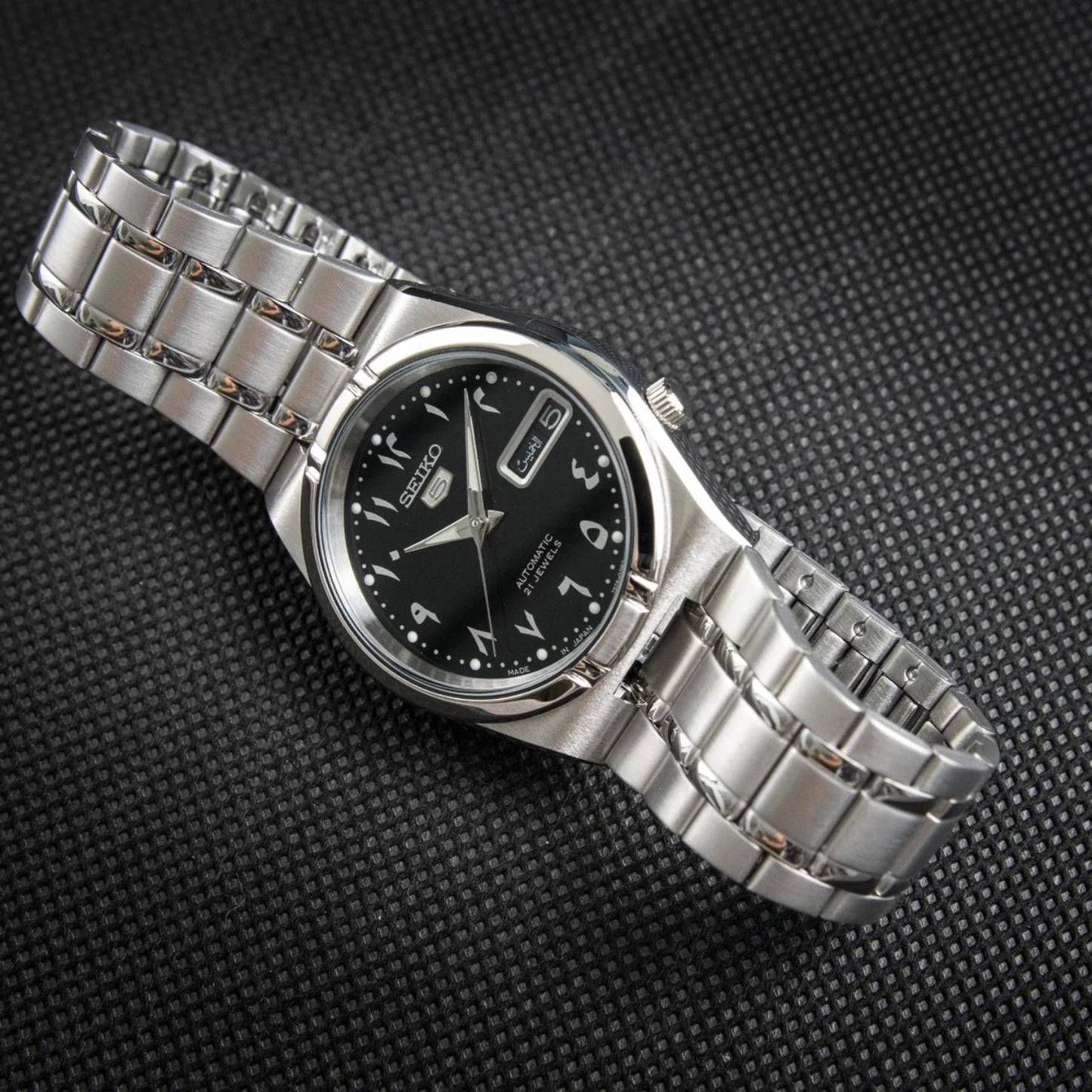 Seiko 5 SNK063J5 automatic watch displayed on a dark textured surface, featuring a black Arabic numeral dial, day-date display, and stainless steel bracelet.