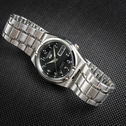 Seiko 5 SNK063J5 automatic watch displayed on a dark textured surface, featuring a black Arabic numeral dial, day-date display, and stainless steel bracelet.