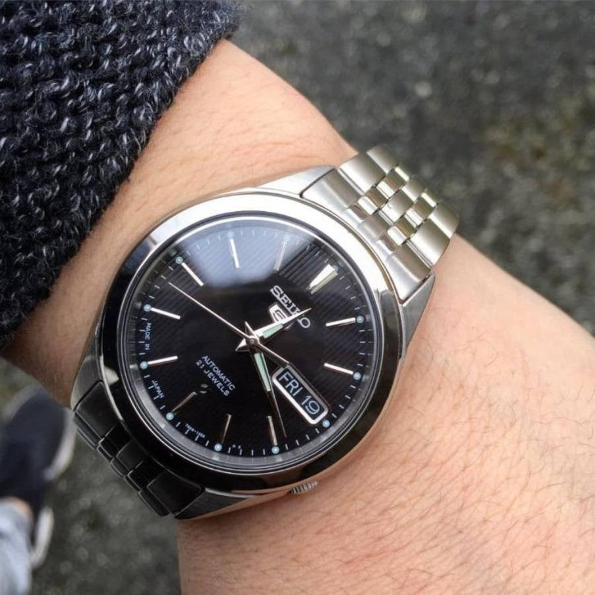 Seiko 5 SNKL23J1 automatic timepiece with a black dial and multi-link stainless steel bracelet displayed on wrist