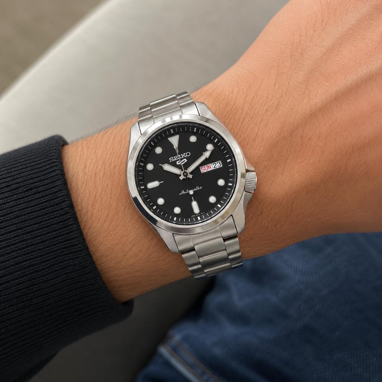 Seiko 5 Sports SRPE55K1 worn on the wrist with black dial, luminous hour markers, day-date display, side crown, stainless steel case, and stainless steel bracelet.