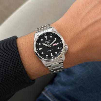 Seiko 5 Sports SRPE55K1 worn on the wrist with black dial, luminous hour markers, day-date display, side crown, stainless steel case, and stainless steel bracelet.