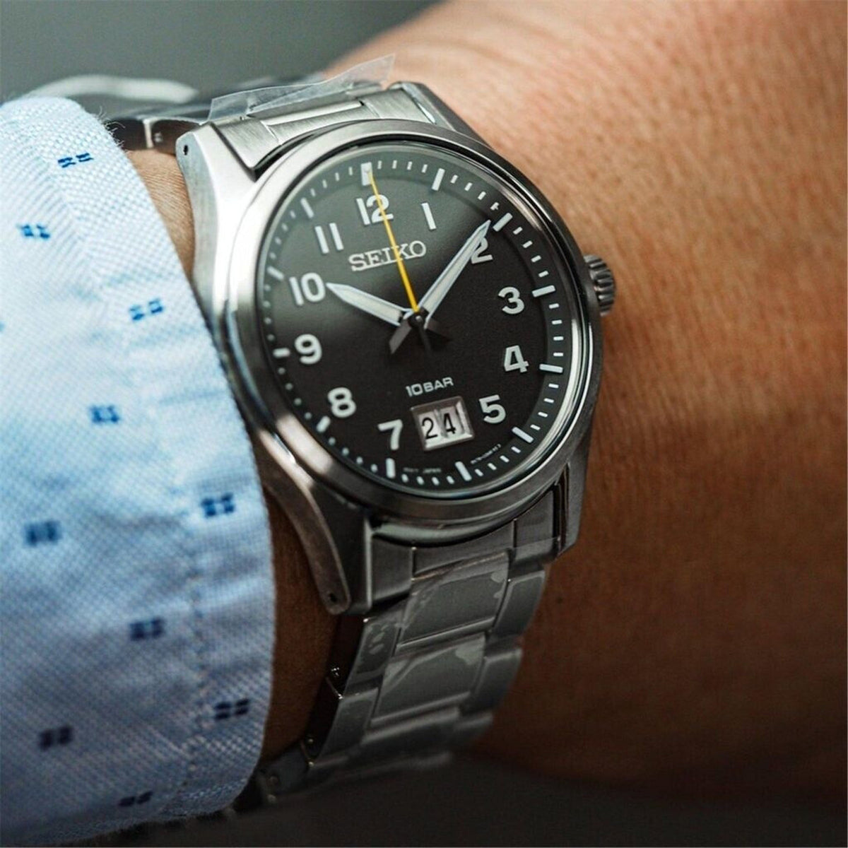 Seiko Classic SUR569P1 men's quartz watch featuring a black dial and steel bracelet worn on the wrist with a blue patterned shirt.