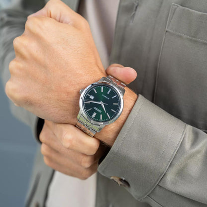 Seiko Essentials SRPH89K1 men's automatic watch worn on the wrist, paired with a green jacket, displaying the green sunburst dial.
