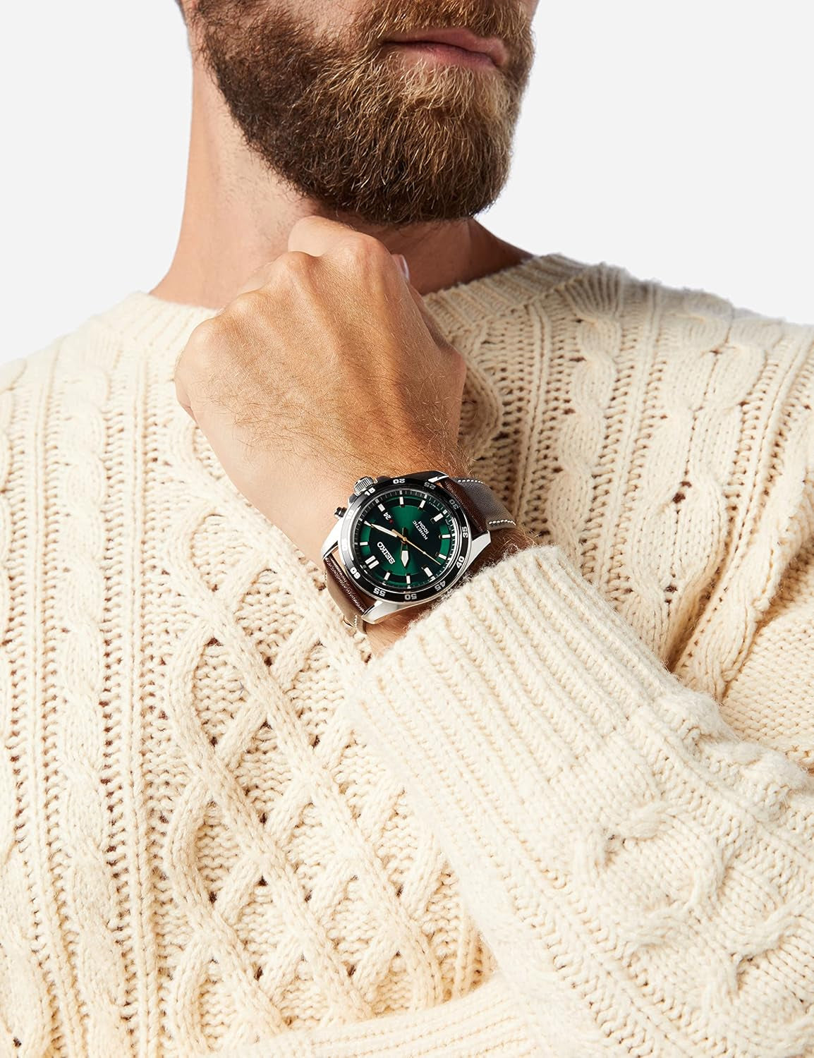 Hombre con jersey beige mostrando en la muñeca un reloj Seiko Kinetic con esfera verde y correa de piel marrón.