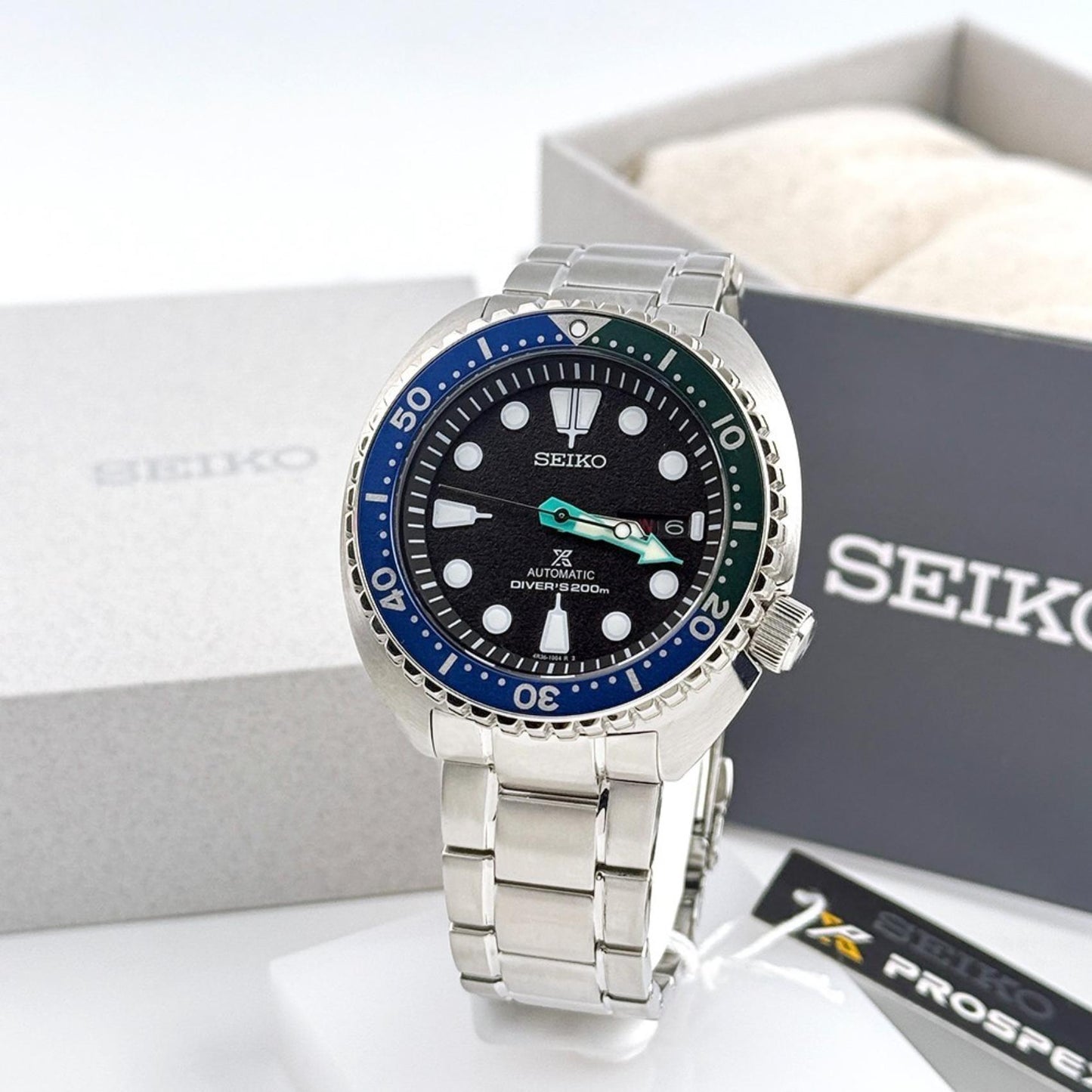 Seiko Prospex SRPJ35 automatic dive watch with black dial, Tropical Lagoon blue-green bezel and stainless steel bracelet displayed beside official Seiko Prospex packaging.