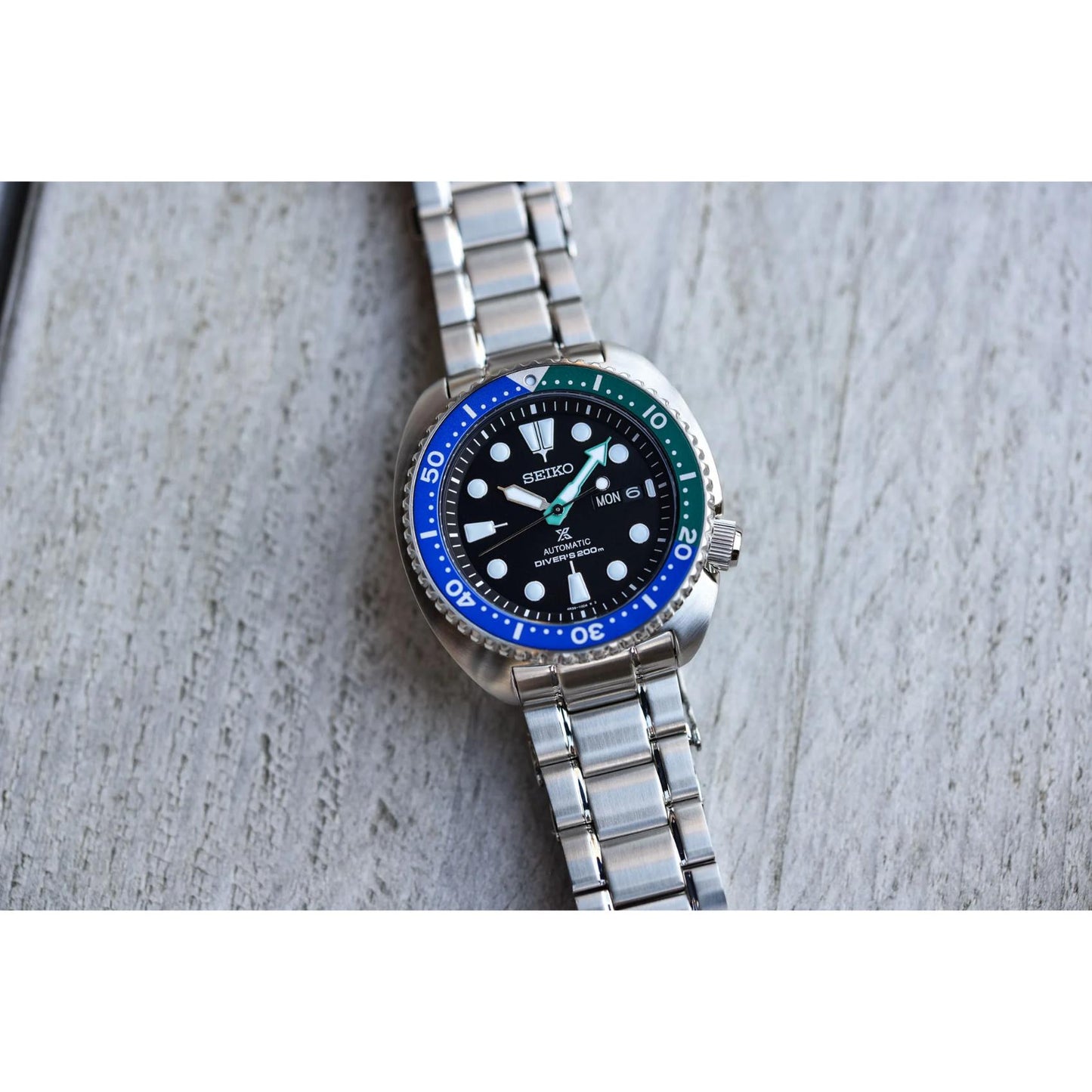 Seiko Prospex SRPJ35 automatic dive watch with black dial, Tropical Lagoon blue-green bezel and stainless steel bracelet displayed on textured wooden surface.