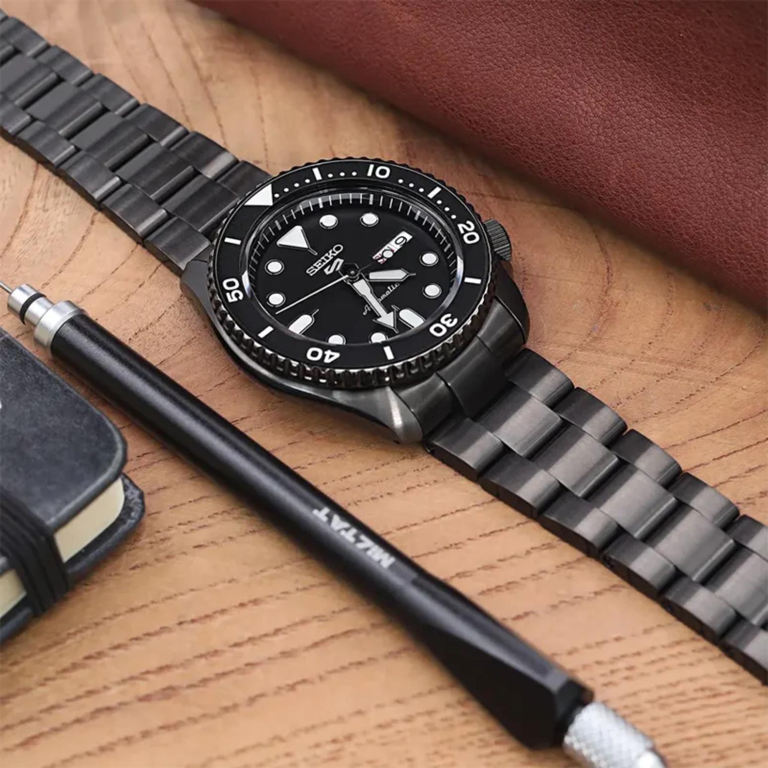 Seiko SRPD65K1 mens automatic dive watch with black dial and bracelet displayed on wooden desk with everyday accessories in lifestyle flat lay
