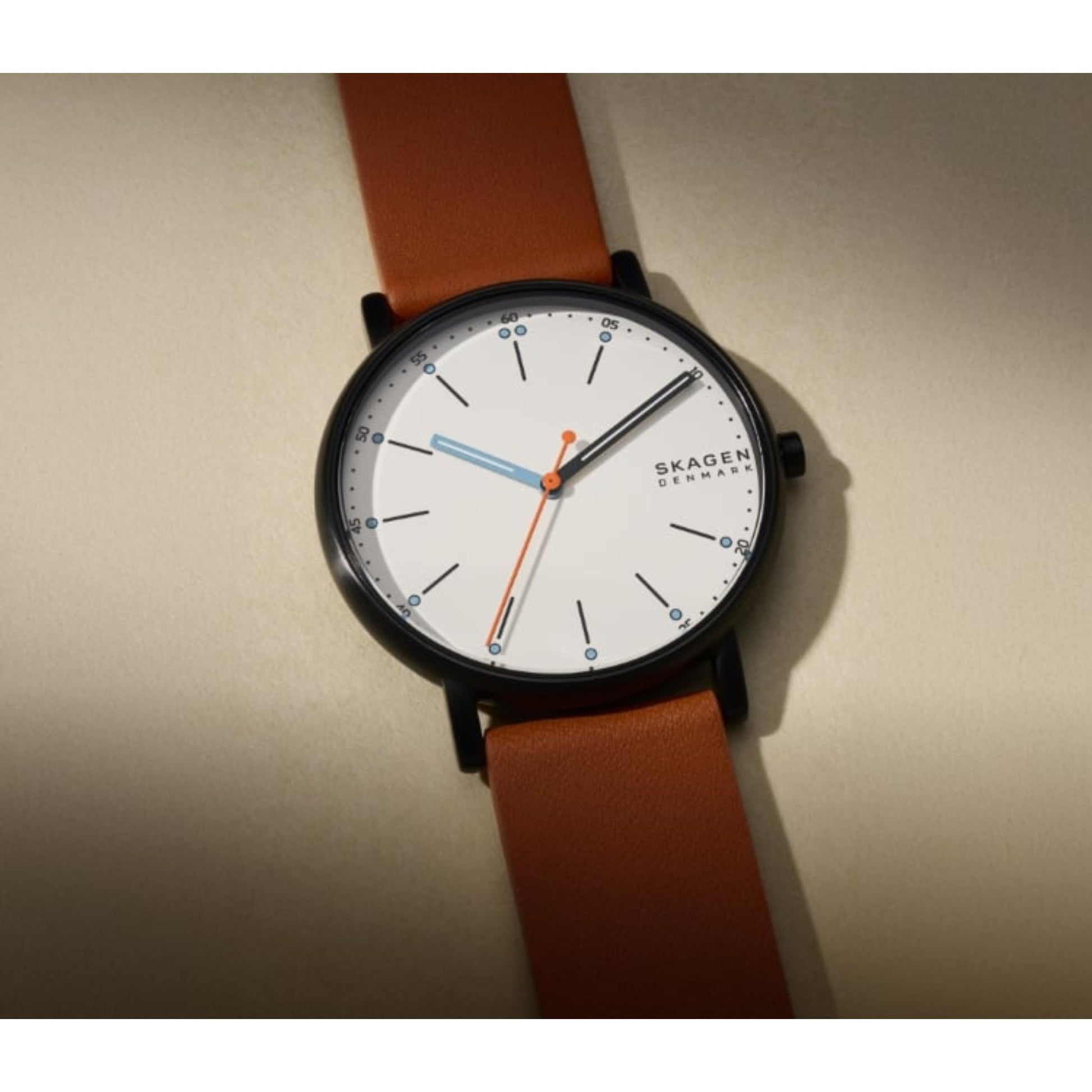 Skagen SKW6374 with white minimalist dial, gunmetal-tone case, orange seconds hand, and brown leather strap shown in a close-up angled view.
