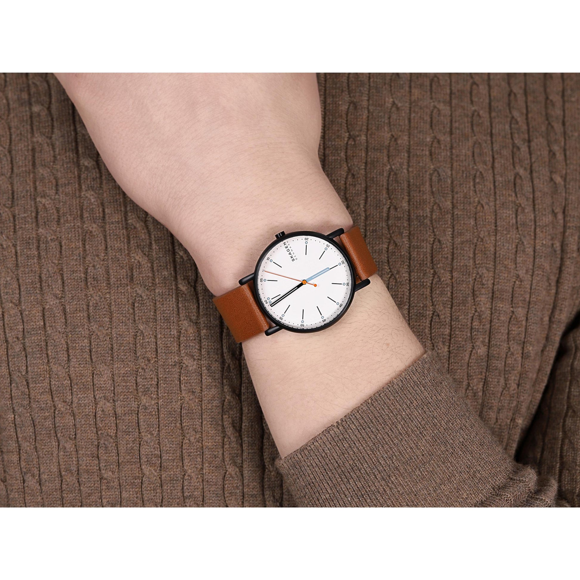 Skagen SKW6374 with white minimalist dial, gunmetal-tone case, orange seconds hand, and brown leather strap worn on the wrist in a casual style.